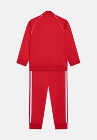 Chándal atlético rojo hecho de tejido suave. Chaqueta con cuello y puños acanalados, pantalones con franjas laterales blancas, ambos tienen un corte relajado.