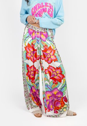 Woman wearing wide-leg pants with large colorful red, pink, and purple floral print, paired with a light blue long-sleeve shirt and open-toe sandals.