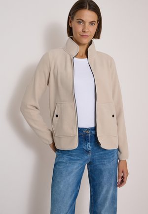 Femme portant une veste beige clair zippée sur une chemise blanche et un jean bleu, debout devant un fond clair uni.