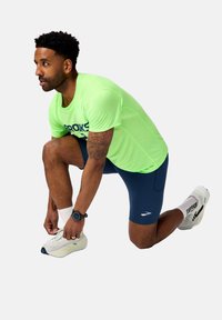 Camiseta atlética verde brillante con el logotipo negro de "Brooks", pantalones cortos azul marino, zapatillas blancas. Persona de rodillas, atándose los zapatos, con un reloj.