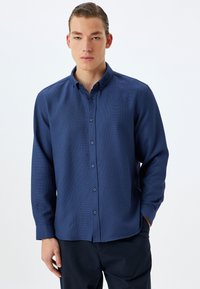 Camisa azul marino de botones con mangas largas, cuello, tejido texturizado y botones a juego, combinada con pantalones oscuros.