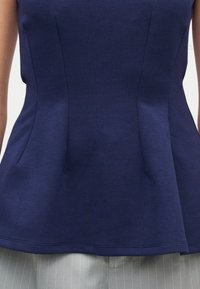 Ναυτικό μπλε εφαρμοστό αμάνικο τοπ με ραφές, φορεμένο πάνω από ανοιχτό γκρι παντελόνι με λεπτές ρίγες.