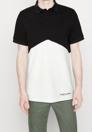 Homme portant un polo bicolore noir et blanc à manches courtes et un pantalon vert, debout devant un fond uni.