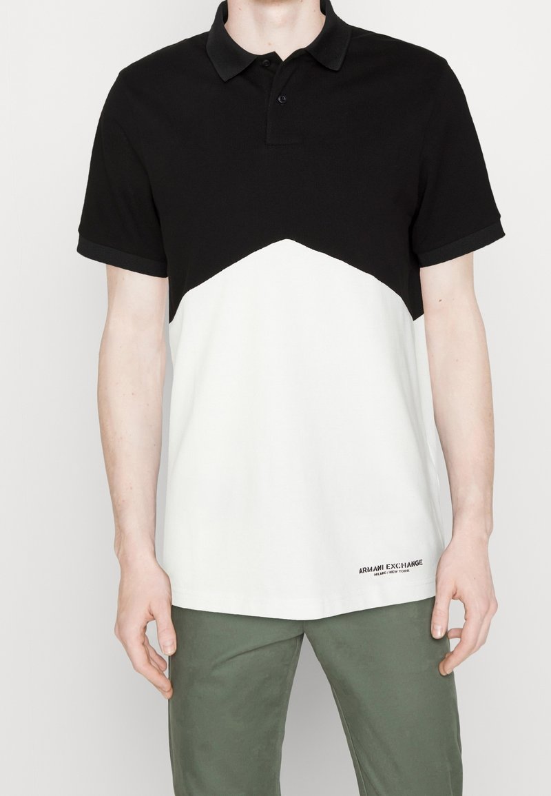 Homme portant un polo bicolore noir et blanc à manches courtes et un pantalon vert, debout devant un fond uni.