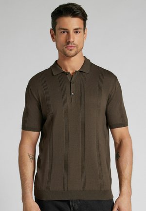 Junger Mann mit kurzen dunklen Haaren, der ein braunes, kurzärmeliges Strick-Poloshirt mit vertikalen, strukturierten Streifen trägt.