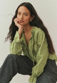 Jasnozielona, teksturowana bluzka z postrzępionymi krawędziami i szerokimi rękawami, zestawiona z ciemnymi, wzorzystymi spodniami. Gładka tkanina o luźnym kroju.