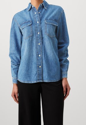 Camisa de mezclilla abotonada con bolsillos con solapa en el pecho, usada con pantalones negros, mostrada desde el cuello hasta las caderas sobre un fondo liso.