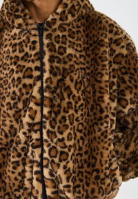 Prim-plan al unei persoane îmbrăcate într-o jachetă din blană sintetică cu imprimeu de leopard maro și negru, cu un fermoar negru, evidențiind detaliile texturii și ale modelului.