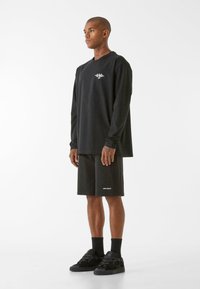 Crna majica dugih rukava s logom na prsima, u kombinaciji s crnim šortsem koji ima bijeli naziv marke. Model stoji u crnim tenisicama i čarapama.