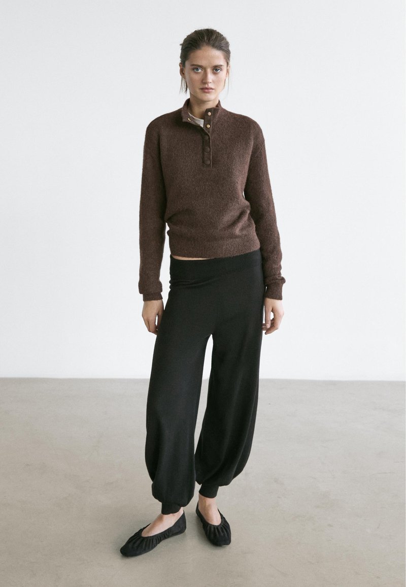 Jeune femme debout contre un mur blanc, portant un pull marron, un pantalon noir ample et des chaussures plates noires.