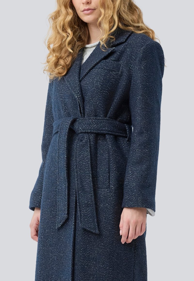Frau mit lockigem blonden Haar, die einen marineblauen, taillierten Wollmantel mit breiten Revers über einem hellgrauen Oberteil trägt.