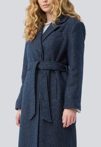 Mujer con cabello rizado rubio que lleva un abrigo de lana azul marino con cinturón y solapas, de pie contra un fondo gris claro liso.