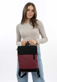 Un sac à dos bordeaux et noir avec un rabat, des bretelles réglables et une texture lisse. Fabriqué en matériau durable, il dispose d'une fermeture éclair sur le côté.