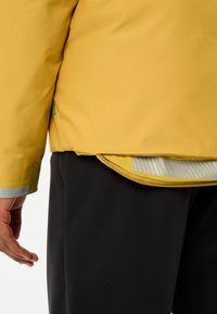 Geel waterdichte jas met een gladde textuur, voorzien van een rits en reflecterende accenten, gecombineerd met zwarte broek.