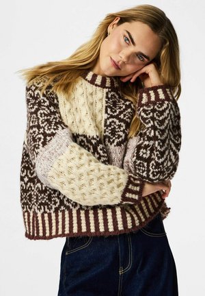 Jeune femme aux cheveux blonds portant un pull en grosse maille aux motifs marron et crème, reposant sa tête sur sa main sur un fond blanc.