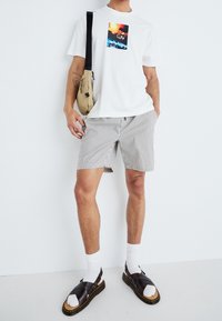 Persona che indossa una maglietta bianca con grafica, pantaloni corti a righe, calze bianche, sandali scuri e porta una borsa a tracolla beige su uno sfondo bianco uniforme.
