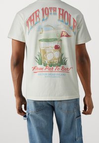 Ljusgrön bomullströja med en grafik av en dryck i ett glas med is och ett körsbär, text som säger "The 19th Hole" och en golfflagga.