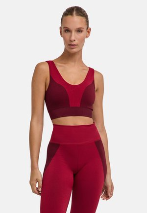 Vrouw draagt donkerrode naadloze sportbeha en bijpassende high-waisted leggings, staand tegen een effen witte achtergrond.