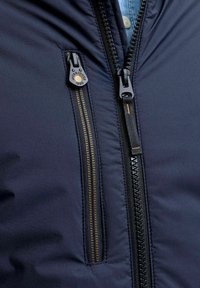 Pomarančna zip-jakna ima teksturirano tkanino s osrednjim zadrgo. Zadrgo krasi zlatno obarvan povodec in kontrastna črna zadrga.