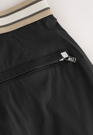 Crne hlačice s teksturiranim pojasom s bež i crnim prugama. Imaju mali džep s patentnim zatvaračem i srebrnim kopčama.