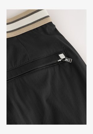 Crne hlačice s teksturiranim pojasom s bež i crnim prugama. Imaju mali džep s patentnim zatvaračem i srebrnim kopčama.