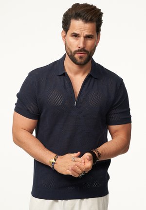 Navyblaues Strickpoloshirt mit geometrischem offenen Muster, kurzen Ärmeln und Halbreißverschlusskragen. Leichter Stoff und strukturierte Oberfläche.