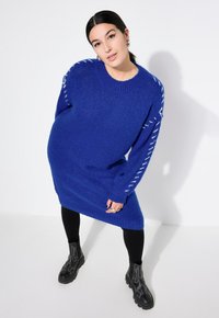 Lys blå strikket sweaterkjole med ribbede kanter og hvide stingdetaljer på ærmerne, parret med sorte tætsiddende leggings og ankelstøvler.