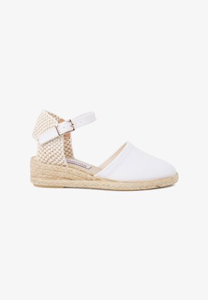 White canvas espadrille with a woven back and ankle strap, featuring a jute wedge sole. Designed with a closed toe and open heel.