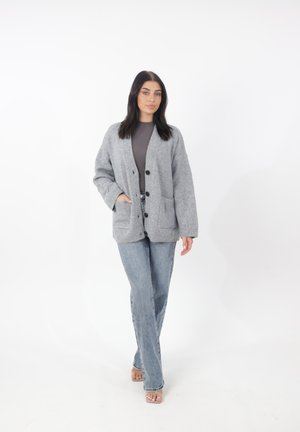 Cardigan en tricot gris à coupe décontractée, avec deux poches avant et fermeture par boutons. Associé à un jean en denim clair et des sandales à bout ouvert.