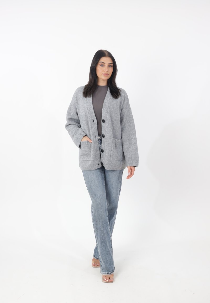 Cardigan grigio lavorato a maglia con vestibilità morbida, due tasche frontali e chiusura con bottoni. Abbinato a jeans di denim chiaro e sandali aperti.