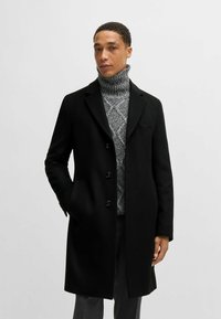 Cappotto lungo in misto lana nero con colletto a punta, cinque bottoni e tasche laterali. Indossato sopra un dolcevita grigio con un motivo a maglia testurizzato.