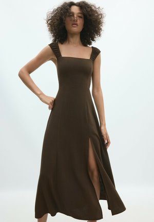 Femme aux cheveux bouclés portant une robe marron sans manches avec des bretelles froncées et une fente haute sur la cuisse, debout devant un fond clair.