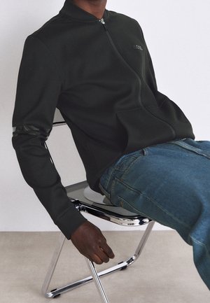 Schwarze Sportjacke mit Reißverschluss aus glattem Stoff, Stehkragen, tonalen Logodetails und gerippten Bündchen, kombiniert mit blauen Jeans.
