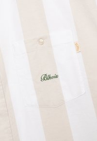 Striped short-sleeve shirt in beige and white, featuring a front pocket with green embroidered text "Blkvis" and a single beige button.