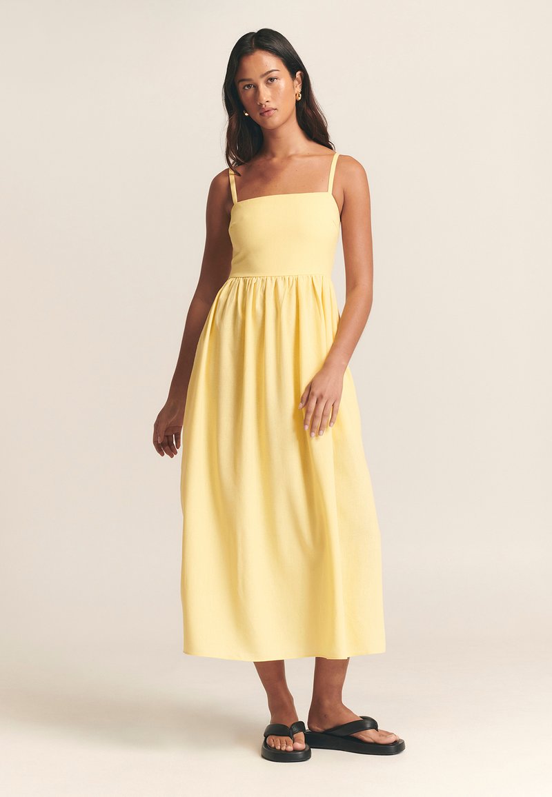 Keltainen midi-mekko, jossa neliömäinen pääntie, ohuet olkaimet ja vyötärö kokoontaitettu. Yhdistetty mustiin tasapohjaisiin sandaaleihin. Sileä kangaspinta.