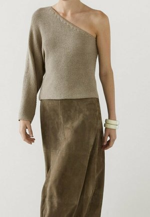 Femme portant un pull en maille taupe à une épaule avec une manche longue et une jupe en daim marron, accessoirisée de deux bracelets blancs au poignet gauche.