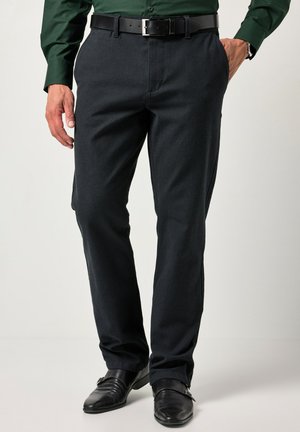 Mann mit dunkelgrauer Hose, schwarzem Ledergürtel, schwarzen Anzugschuhen und einem dunkelgrünen Hemd mit Knöpfen, steht mit einer Hand in der Tasche.