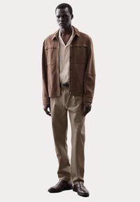 Chaqueta de ante marrón sobre una camisa beige clara, combinada con pantalones caqui y zapatos de cuero marrón. Actitud autoritaria, diseño minimalista.