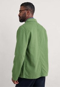 Green canvas jacket with a straight cut, featuring a pointed collar and side pockets, shot from the back, showing textured fabric.