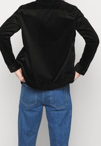 Camicia in velluto a coste nera con taglio dritto e polsini abbottonati, indossata sopra jeans in denim blu. Tessuto testurizzato con righe visibili.