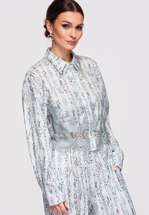 Femme portant un chemisier bleu clair à motifs avec col en strass et partie centrale en dentelle, associé à un pantalon assorti, posant avec la main dans la poche.