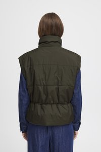 Oliivroheline ilma käisteta puffvest kõrge kraega, kitsendatud taljega ja horisontaalse õmblusega, kantud teksasärgi peal.