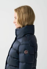 Marineblauwe pufferjack met hoge kraag en doorgestikt ontwerp. Voorzien van een gemarkeerde ronde badge aan de linkerkant. Gladde, glanzende textuur.
