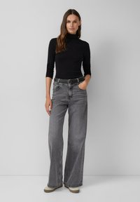 Zwarte coltrui met aansluitende mouwen; grijze high-waisted, wijde jeans met riem; neutraal gekleurde schoenen met een gestructureerde zool; effen achtergrond.