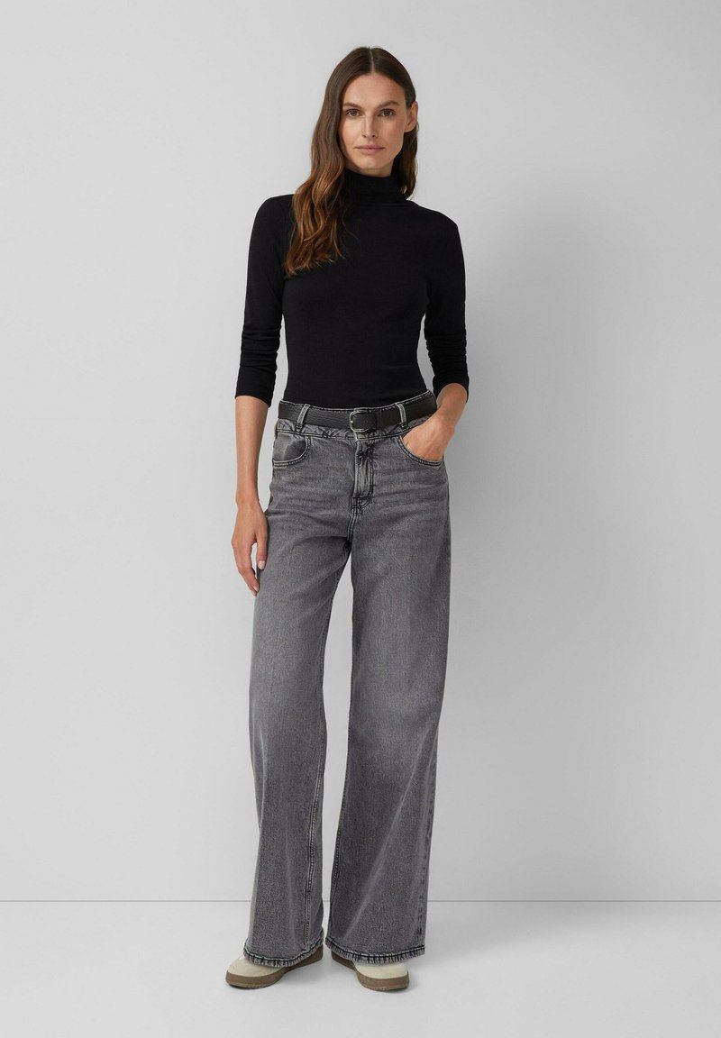 Zwarte coltrui met aansluitende mouwen; grijze high-waisted, wijde jeans met riem; neutraal gekleurde schoenen met een gestructureerde zool; effen achtergrond.