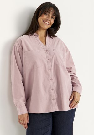 Vrouw glimlachend, draagt een losse lichtroze blouse met knopen en donkere broek, staand tegen een witte muur met hand rustend op de heup.