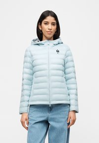 Γυναίκα με ανοιχτό μπλε μπουφάν με κουκούλα και μπλε τζιν παντελόνι, στέκεται μπροστά σε απλό λευκό φόντο.