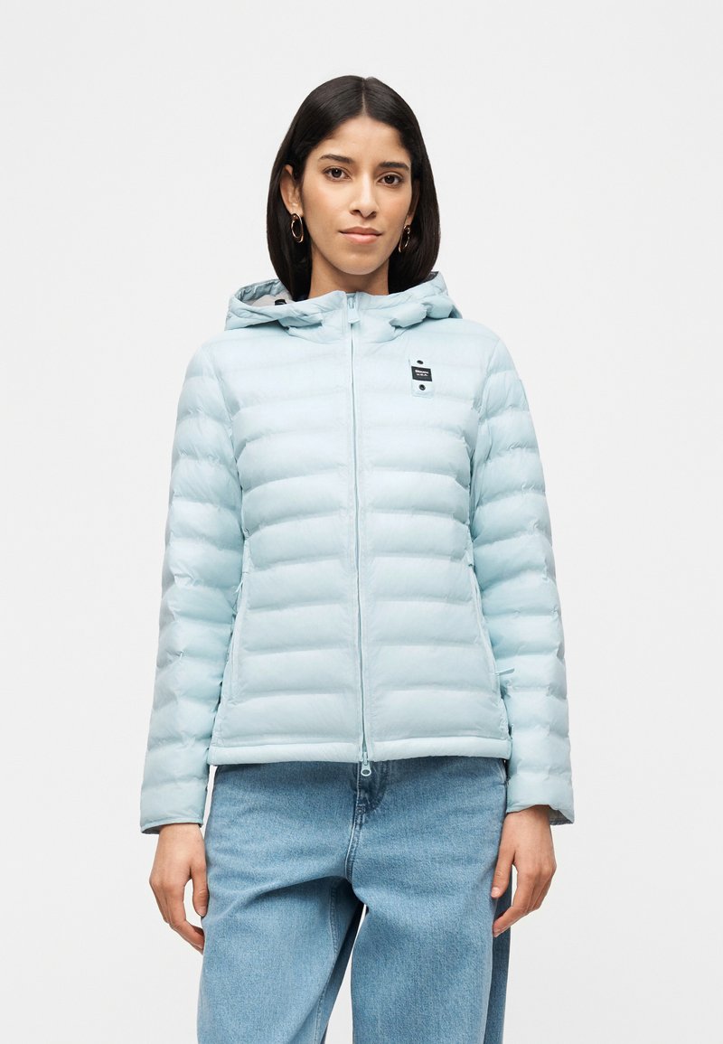 Γυναίκα με ανοιχτό μπλε μπουφάν με κουκούλα και μπλε τζιν παντελόνι, στέκεται μπροστά σε απλό λευκό φόντο.