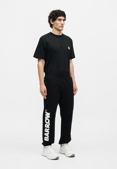 Άνδρας όρθιος, κοιτά προς τα εμπρός φορώντας μαύρο μπλουζάκι με μικρό κίτρινο λογότυπο και μαύρο παντελόνι φόρμας με μεγάλο λευκό κείμενο "BARROW", λευκά sneakers.