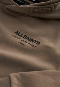 Smeđa dukserica s kapuljačom, s crnim izvezenim natpisom "ALLSAINTS" i akcentima. Glatka tekstura tkanine, minimalizam dizajna.